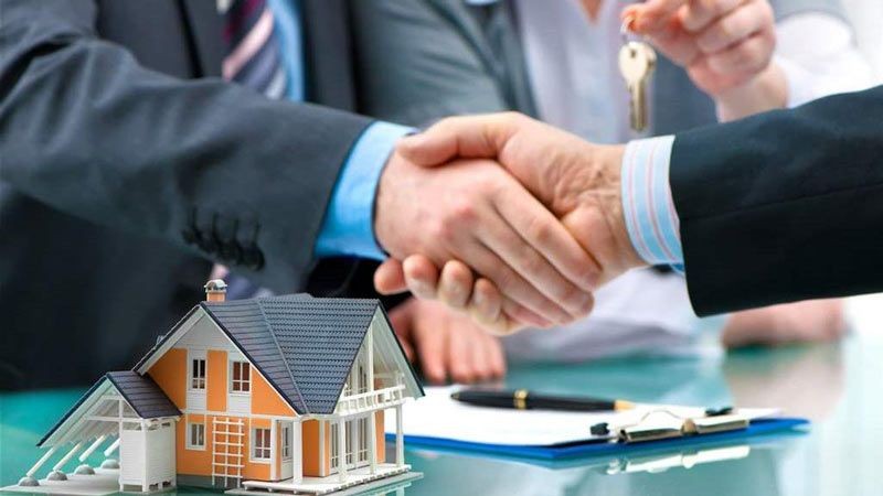 Two individuals shaking hands over a desk with a small model house, keys, and contract documents.