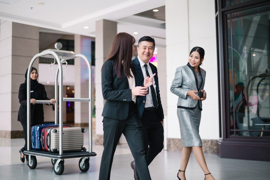People in business attire walking through a hotel lobby with a luggage cart in the background.